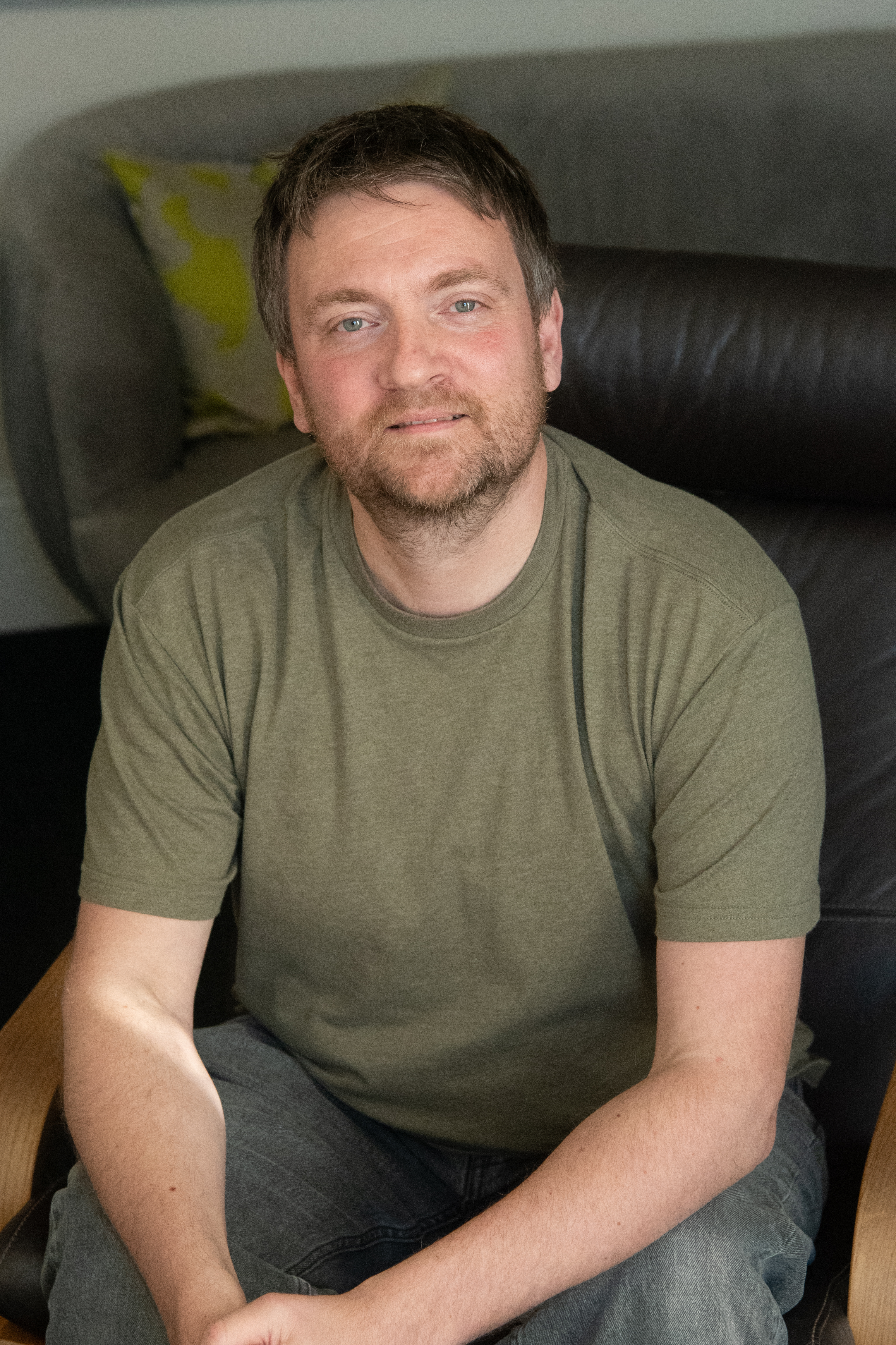 Tim, therapist at Harley Street Counselling, smiling in green t-shirt on grey sofa.